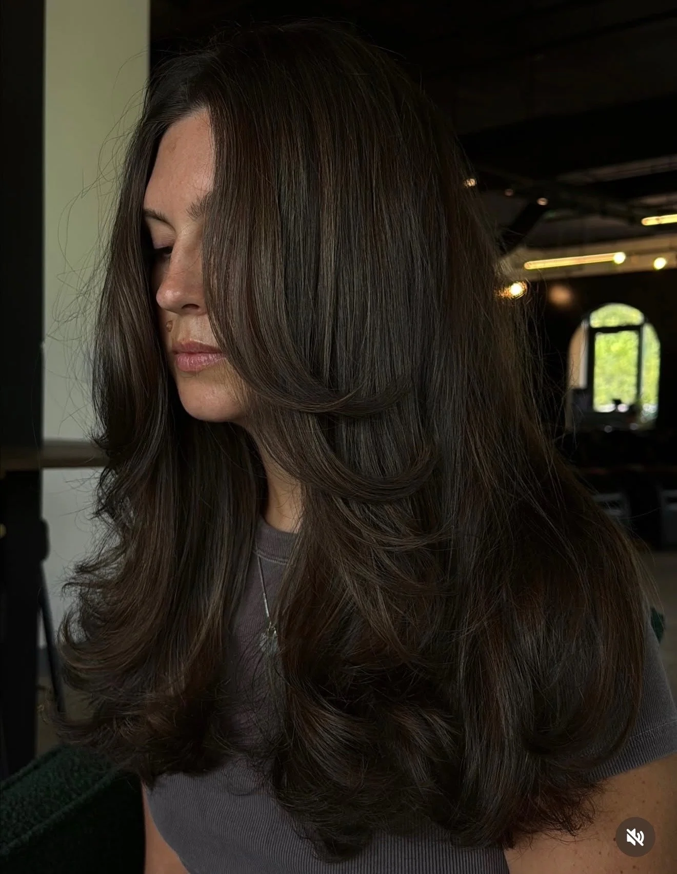A woman with long, wavy dark brown hair and light skin, wearing a gray top, standing indoors with a dark background and light coming from a window.