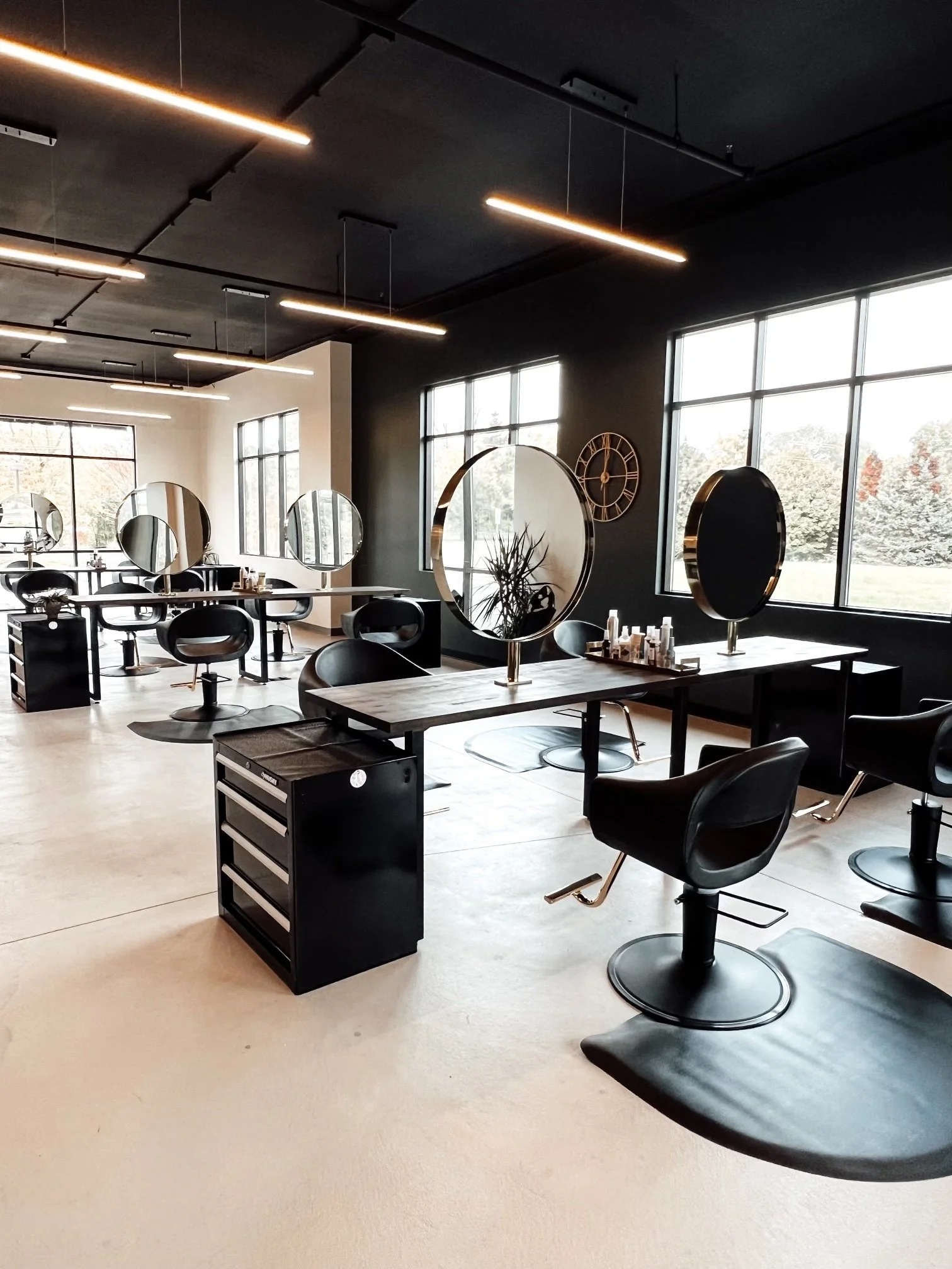 A modern hair salon with black styling chairs, mirrors, and large windows showing a snowy outdoor scene. The salon has black and white walls, a black ceiling with linear lighting, and a clock on the wall.
