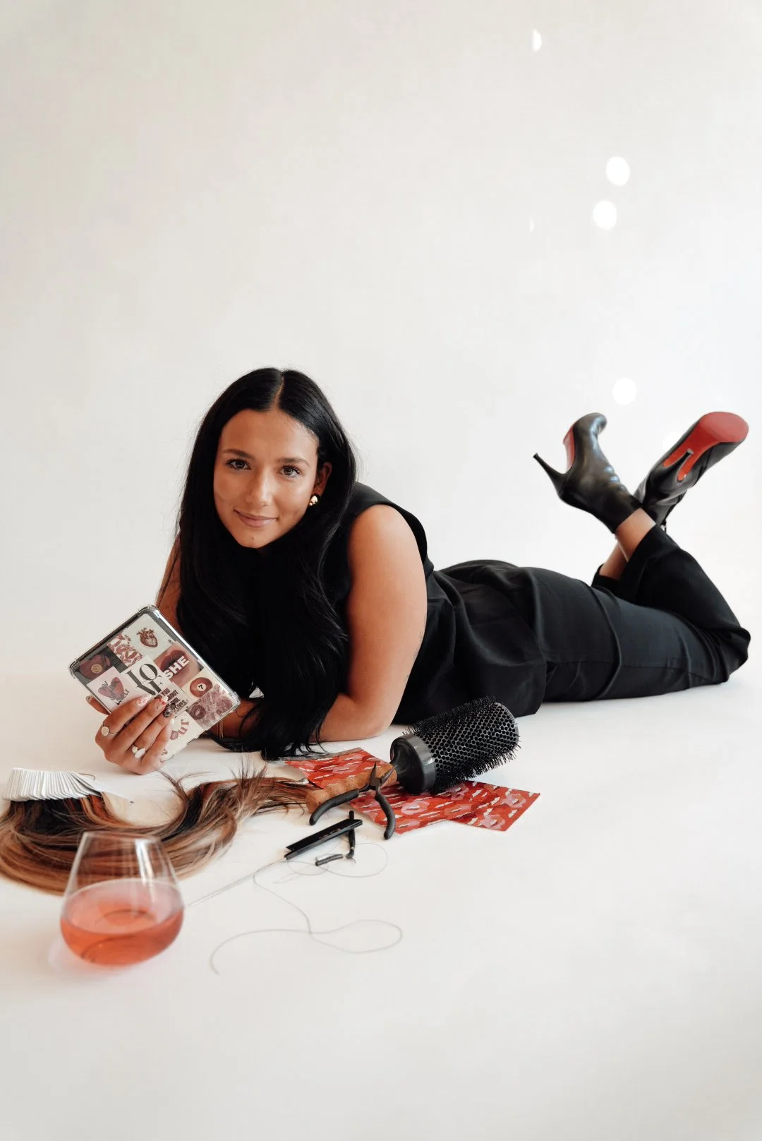Woman with long black hair lying on her stomach on a white floor, holding a makeup palette, surrounded by hair styling tools, a hair wig, a glass of pink drink, and hair color samples.