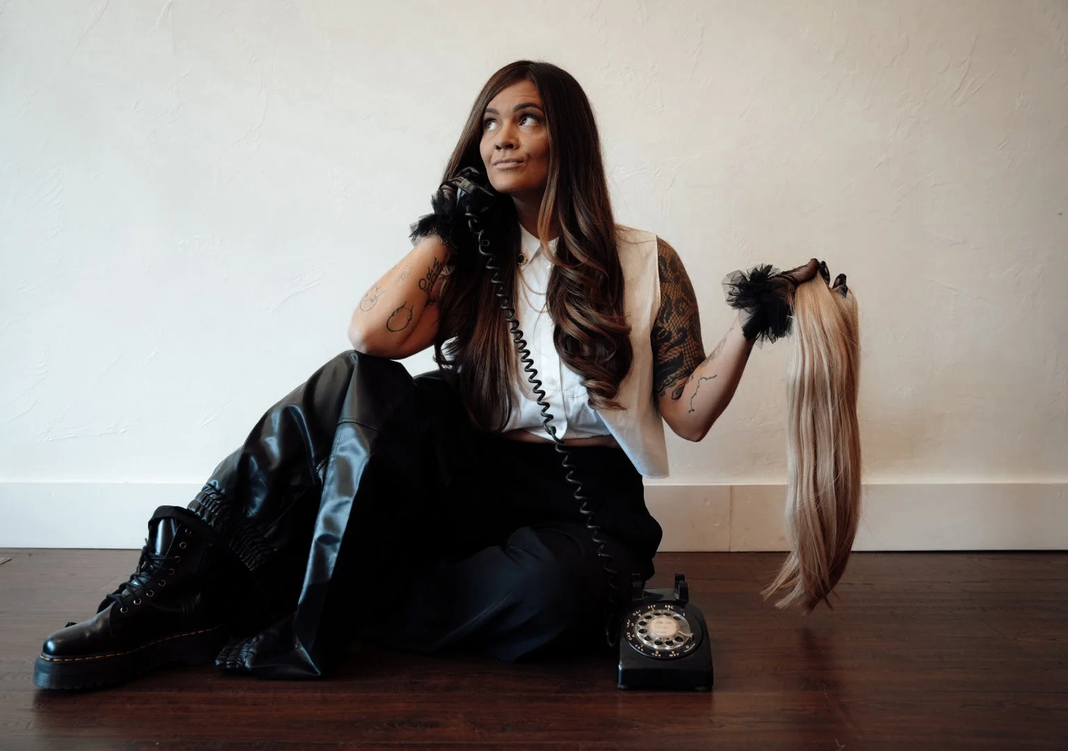 A woman with long brown hair, tattoos, and black gloves, sitting on the floor with a vintage rotary phone, holding a blonde wig, and wearing black boots and pants, against a plain white wall.
