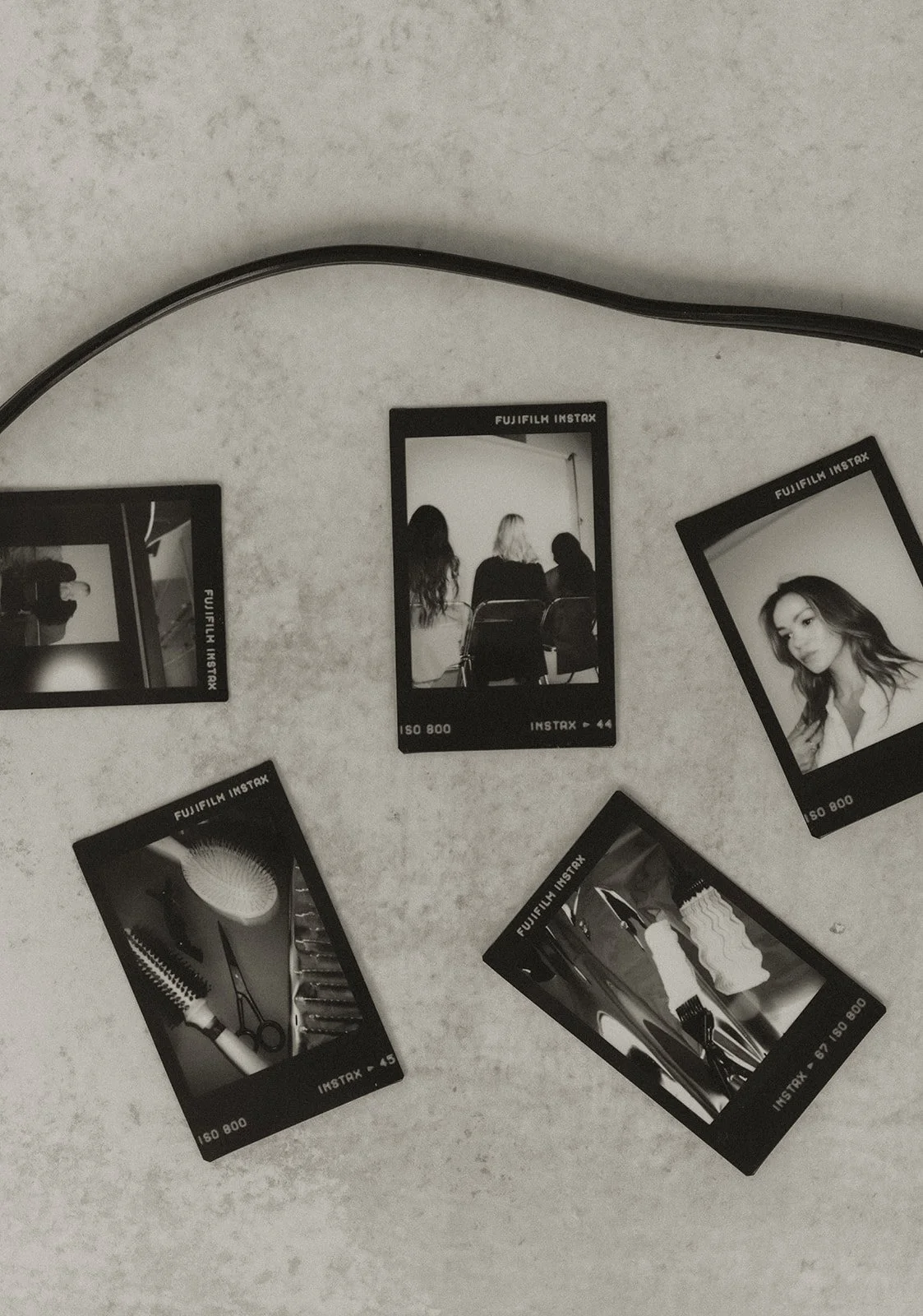 Black and white instant photographs scattered on a light surface, featuring a hairbrush, a woman with wavy hair, a group seated in a room, a woman with long dark hair, and various objects.