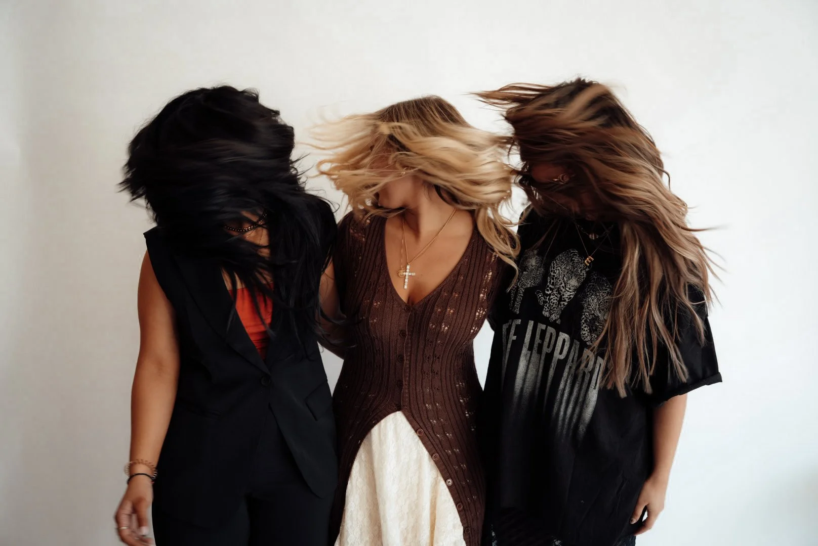 Three women with long hair are standing together, heads tilted downward and hair in motion, against a plain light background.