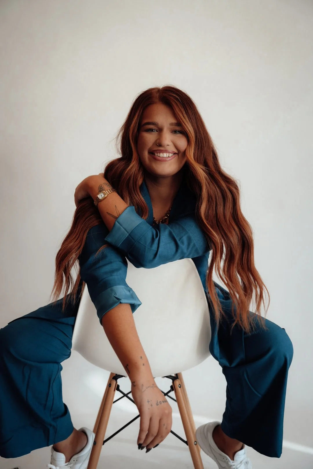 A woman with long, wavy red hair smiling while sitting on a white chair with wooden legs, wearing a blue suit and white sneakers, against a plain white wall.