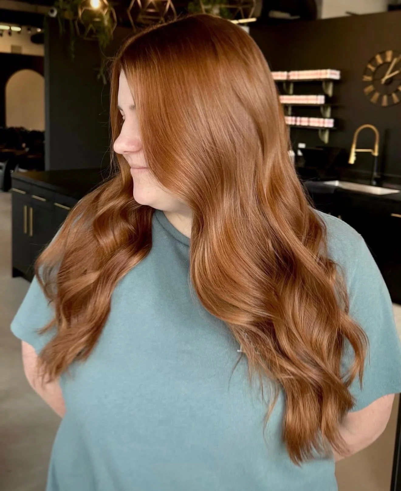 A woman with long wavy red hair wearing a light blue t-shirt standing in a modern kitchen with black cabinets, a gold faucet, and shelves with rolled towels.