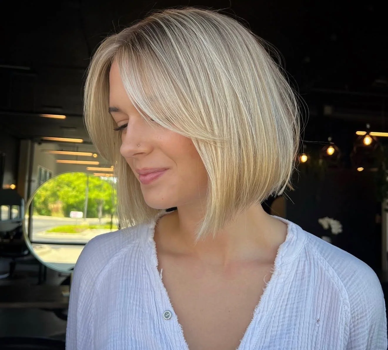 A woman with a short blonde bob hairstyle, wearing a white top, is smiling with her eyes closed inside a modern cafe with warm lighting and large windows showing greenery outside.