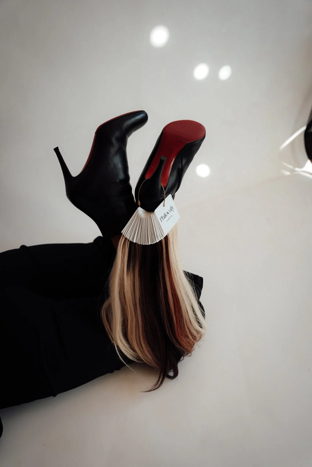 A person holding a black high-heeled shoe with a red sole and a white tag labeled "Philosophy" against a plain white background.