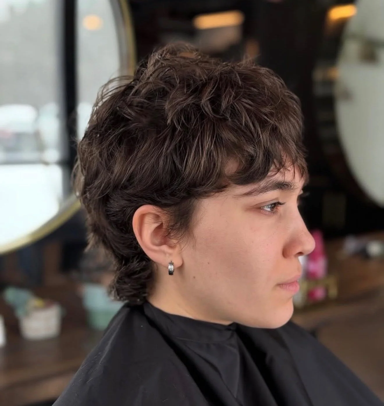 A woman with short, curly brown hair, silver hoop earrings, and a black cape sits in a salon chair in a hair salon, preparing for a haircut or styling.