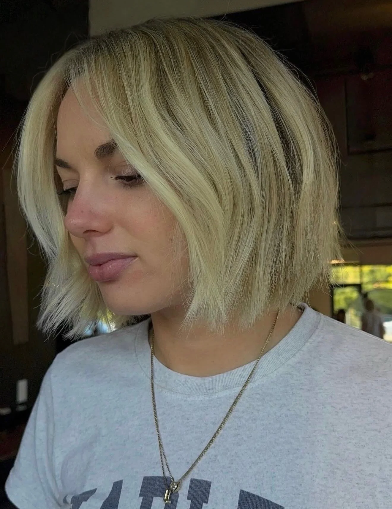 A woman with blonde, shoulder-length hair styled in a bob, wearing a light gray T-shirt and a silver necklace, poses in an indoor setting with natural light.