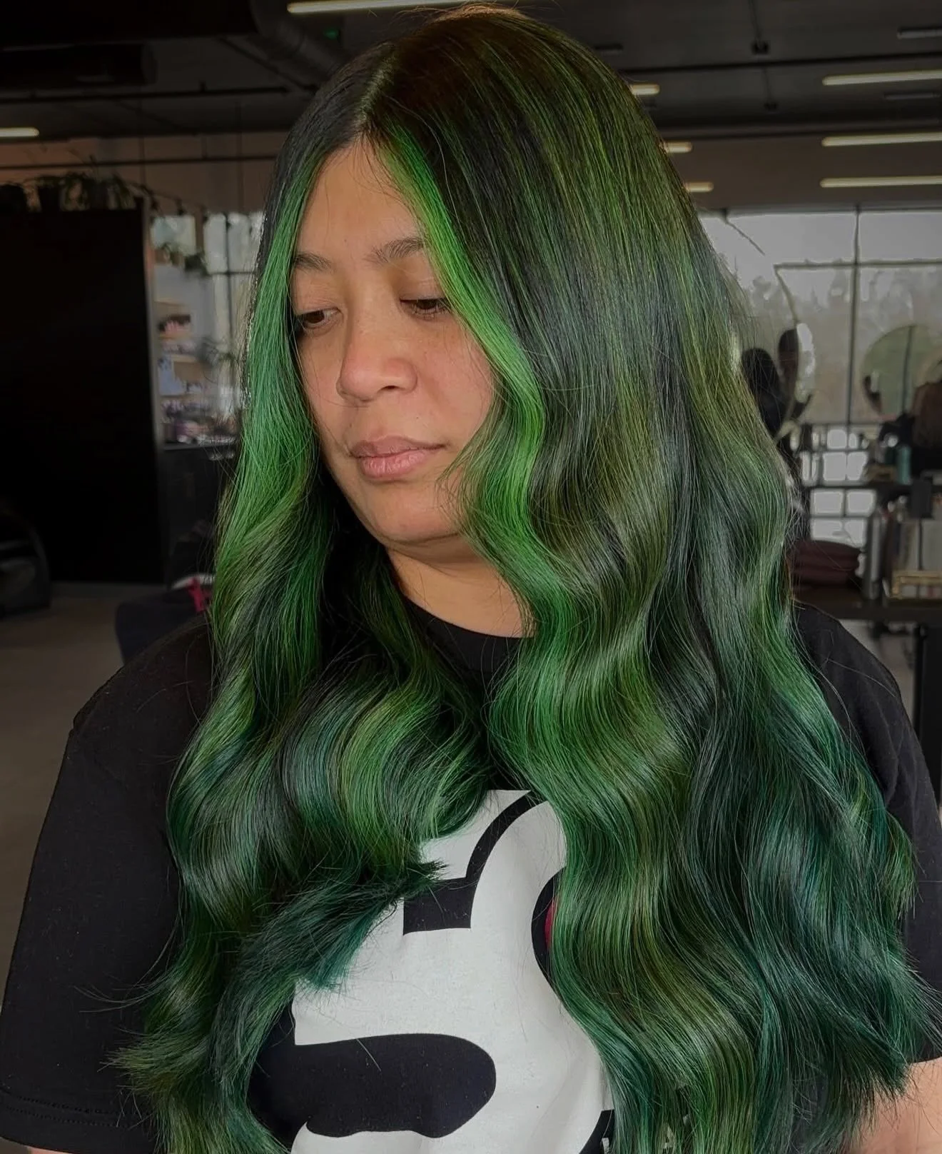 A woman with long wavy black hair accented with vibrant green streaks, standing indoors with a neutral expression.