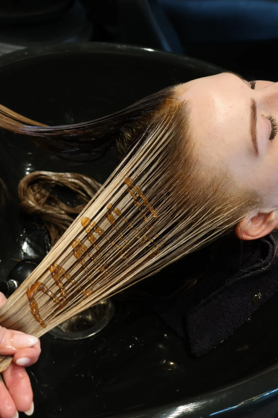 A person with long hair is having their hair washed and treated in a salon, with hair strands extended on a comb showing a treatment process.