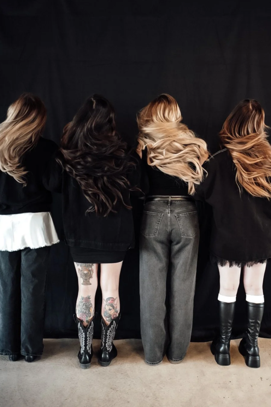 Four women with long, wavy hair, seen from behind, standing against a black background, wearing predominantly black clothing including jeans, skirts, and boots.