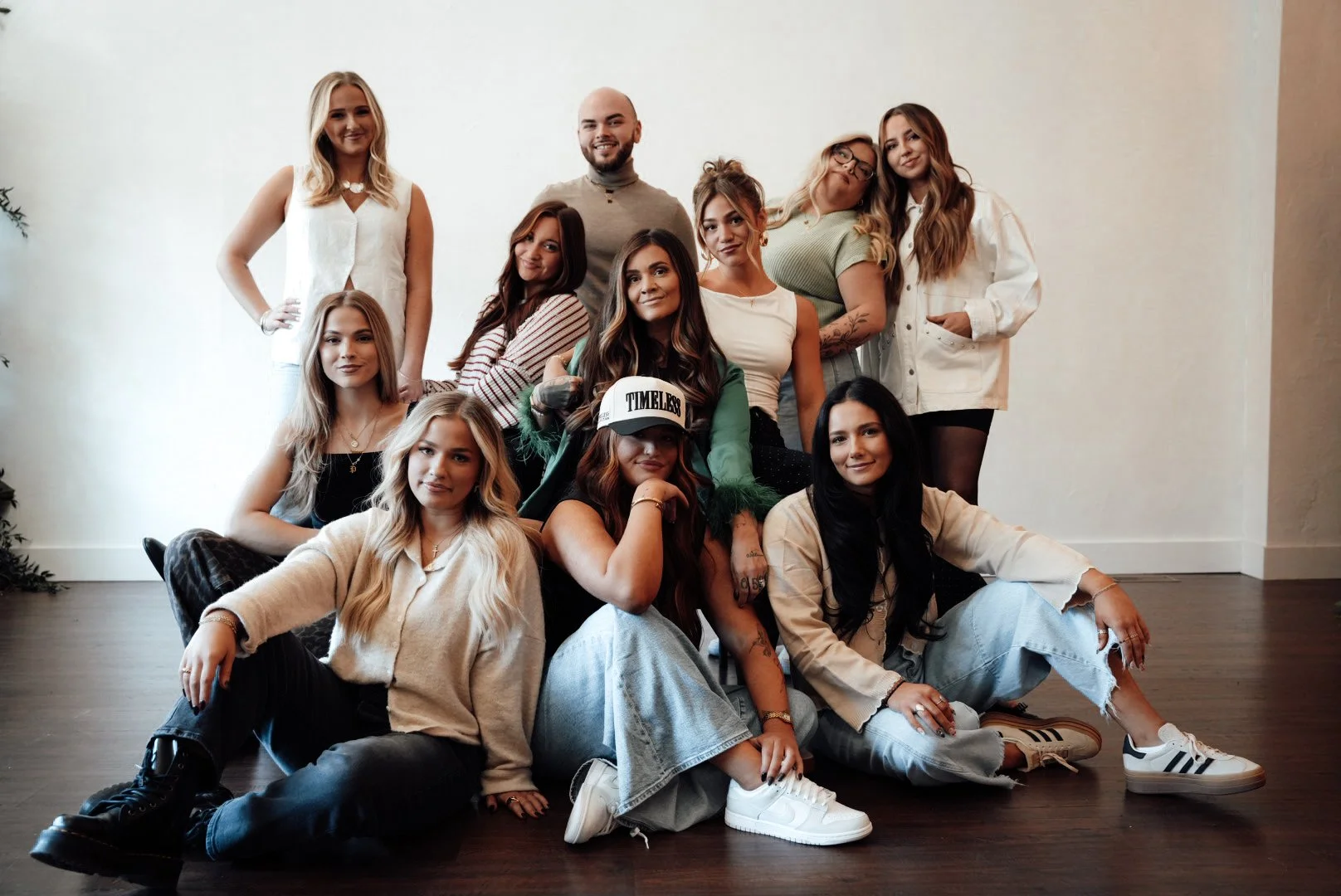 A group of eleven diverse young women and one man posing together indoors. They are dressed casually, some sitting on the floor, others standing or leaning against each other, smiling at the camera.