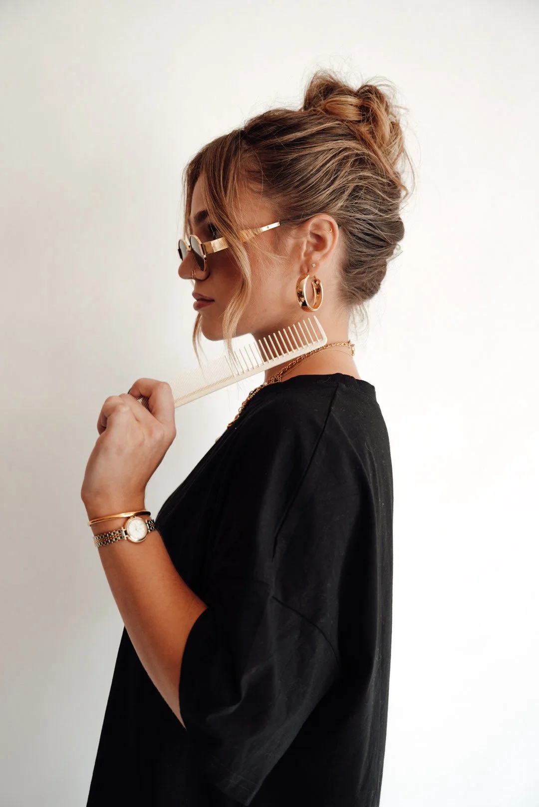 Side profile of a woman with blonde hair styled in a messy bun, wearing sunglasses, gold hoop earrings, a black top, and holding a hair comb to her hair.