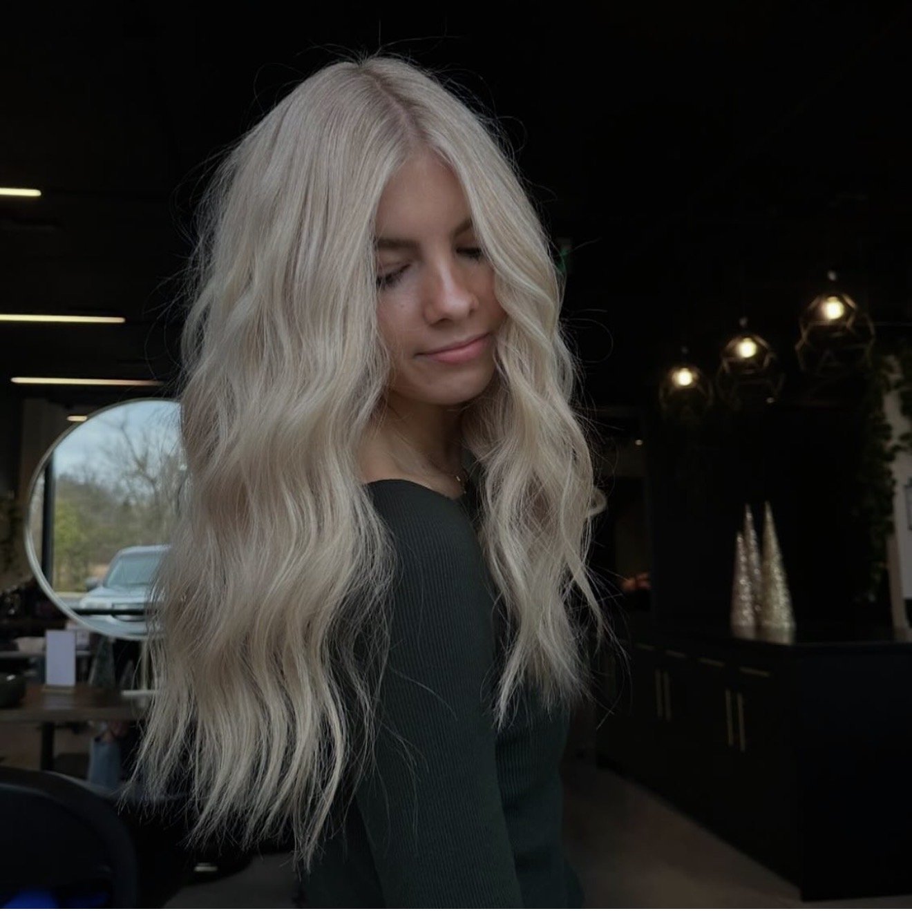 A woman with long wavy blonde hair and closed eyes inside a modern cafe, with tables, chairs, and decorative elements in the background.