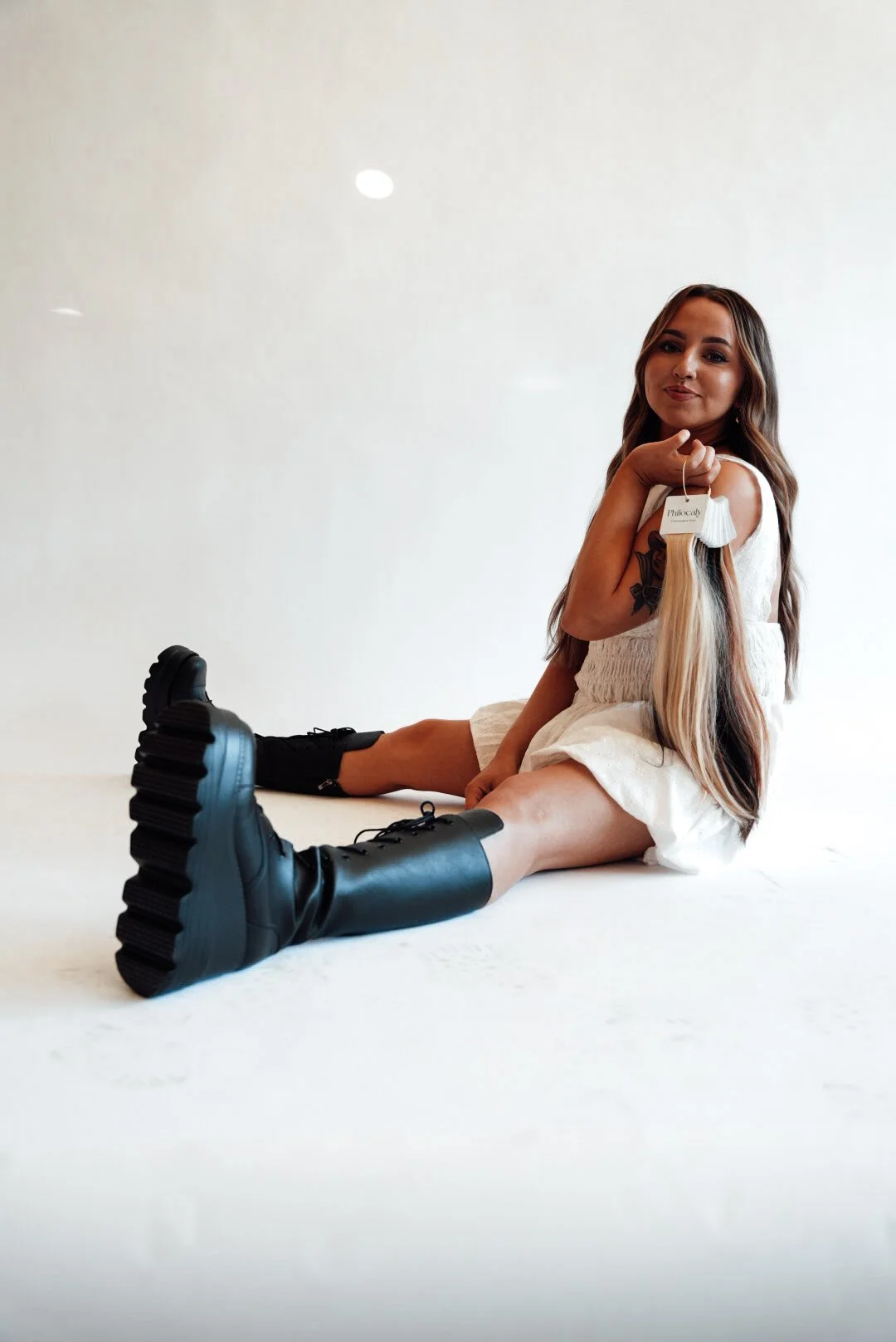 A young woman with long brown hair sitting on a white surface against a white background, wearing black combat boots and a white dress, holding a blonde hair extension with a tag attached.