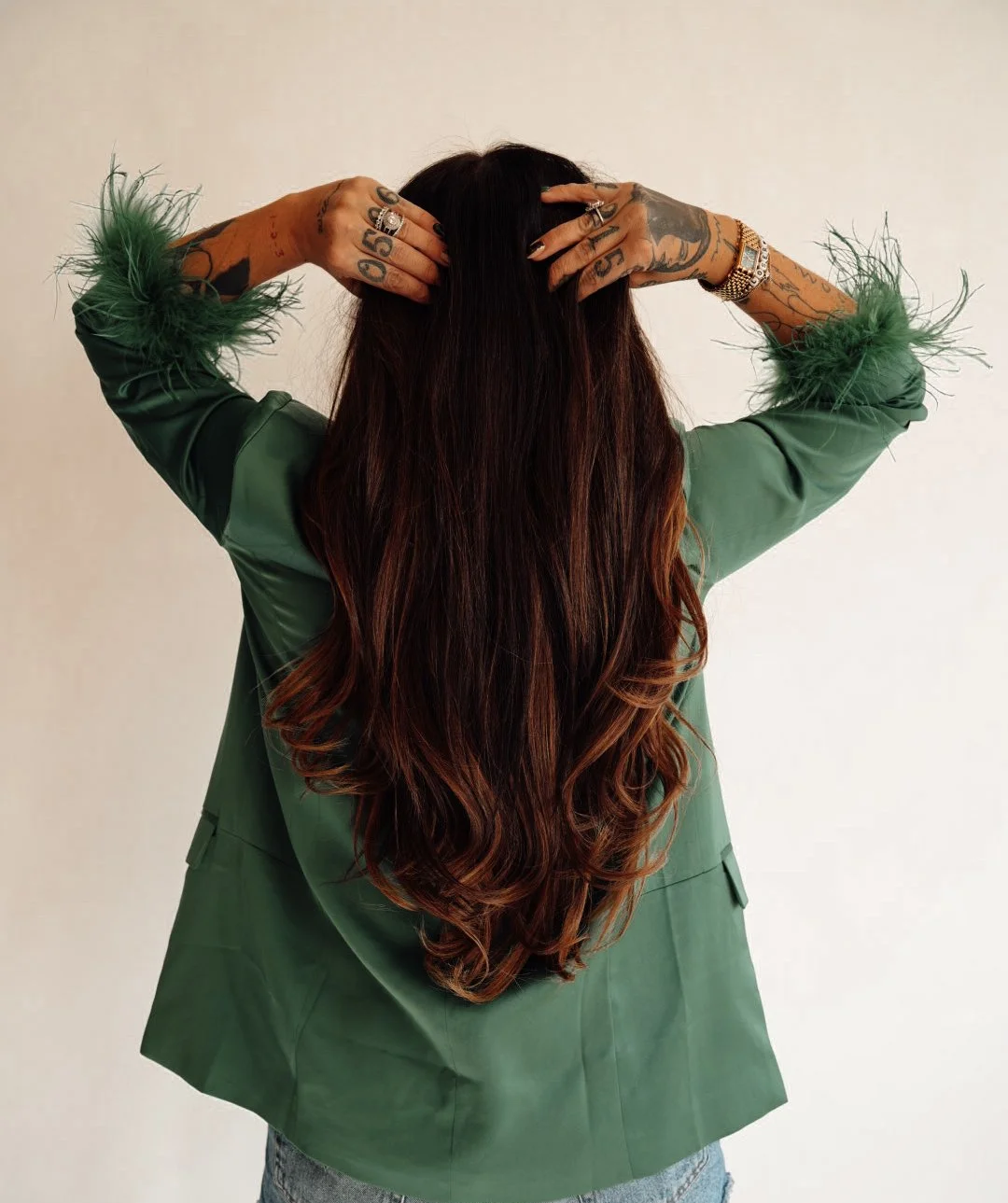 Person with long, wavy brown hair wearing a green jacket with feathered cuffs, facing away, with tattoos on their hands and wearing jewelry, standing against a plain background.