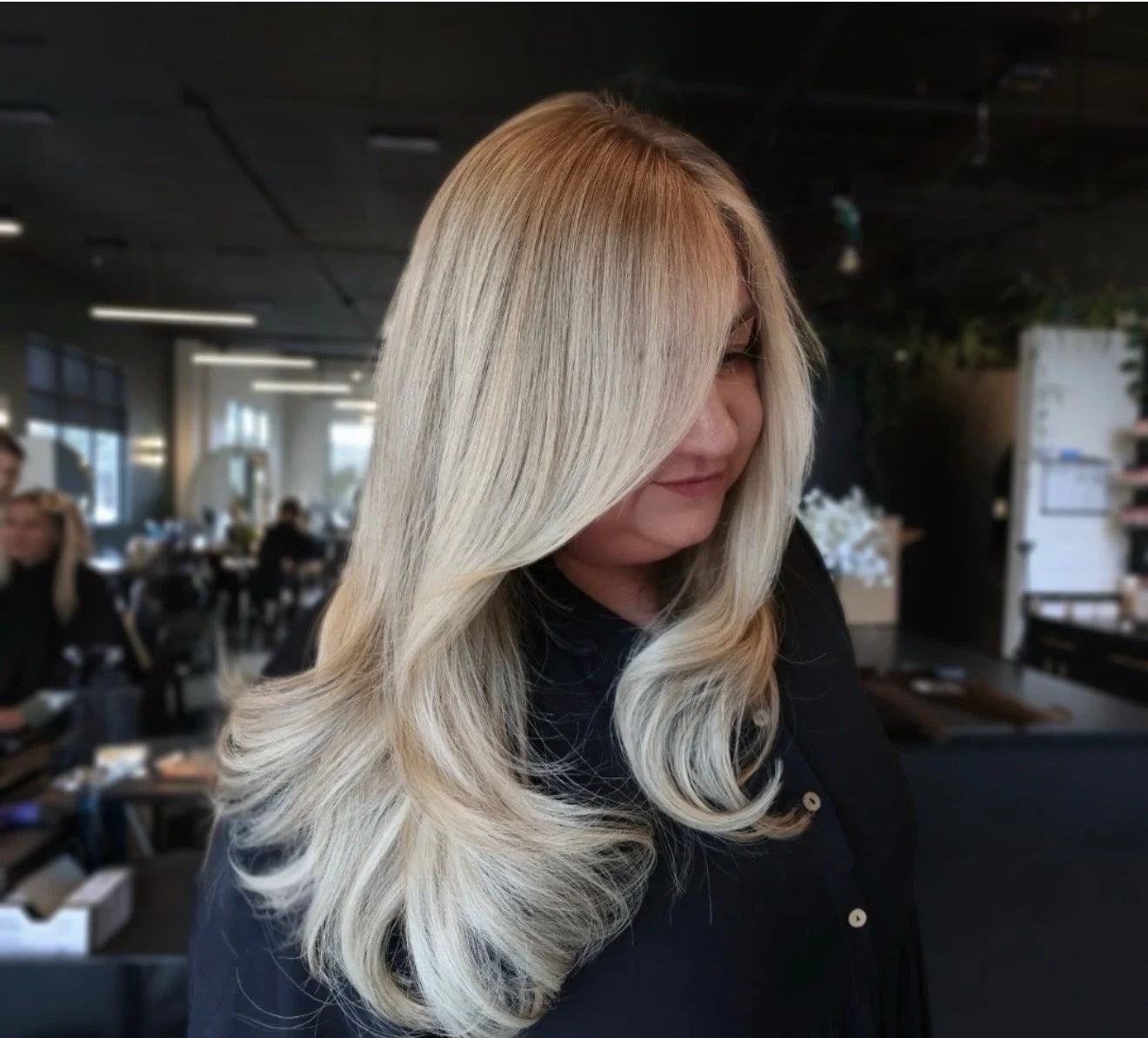 Woman with long, wavy blonde hair with darker roots, inside a salon, with people and salon equipment in the background.