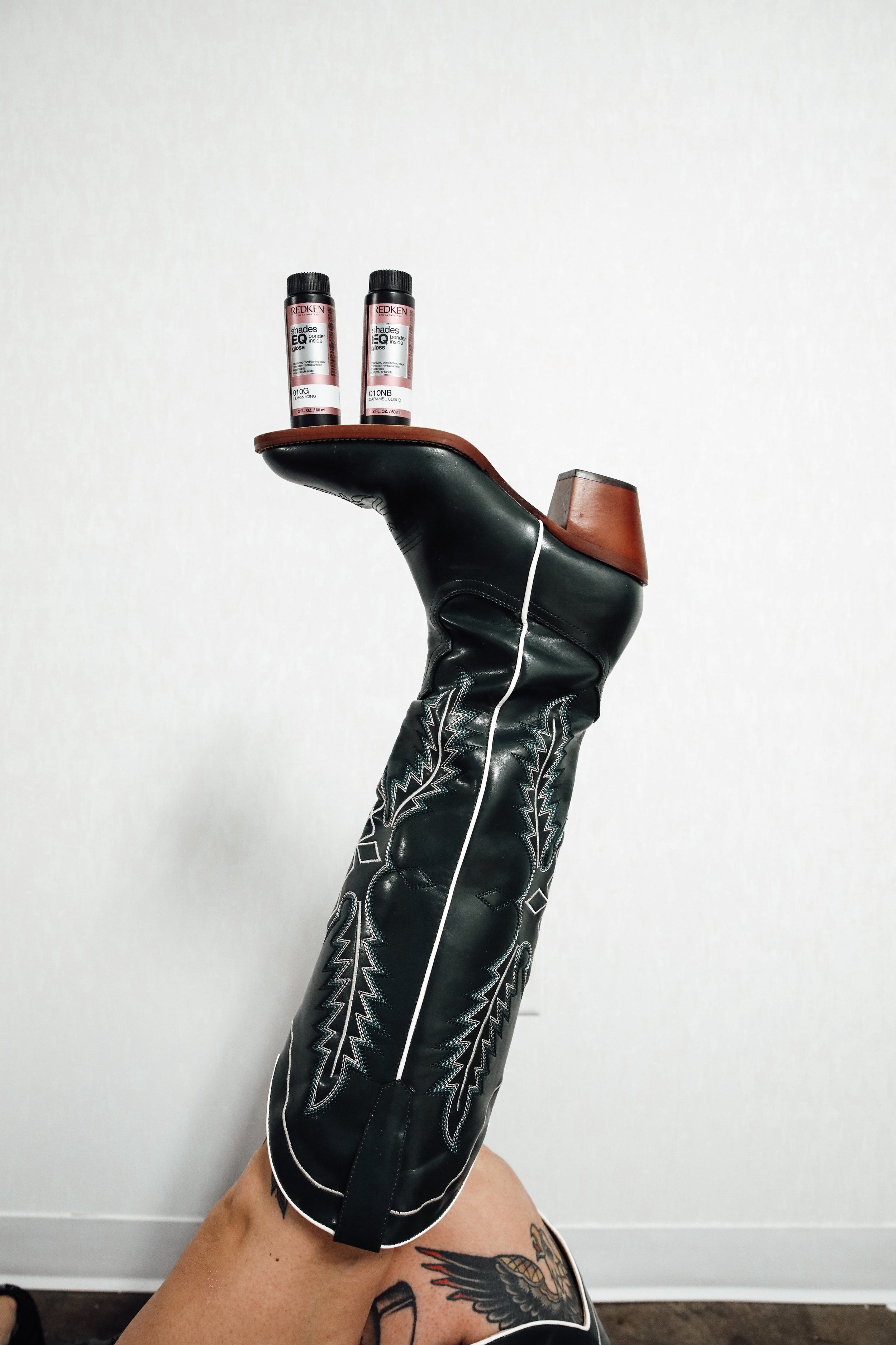 A hand wearing a black leather glove holding two tubes of Redken shades EQ Bonding Gloss, one labeled '010G' and the other '010NB,' with a tattooed leg inside a white room.