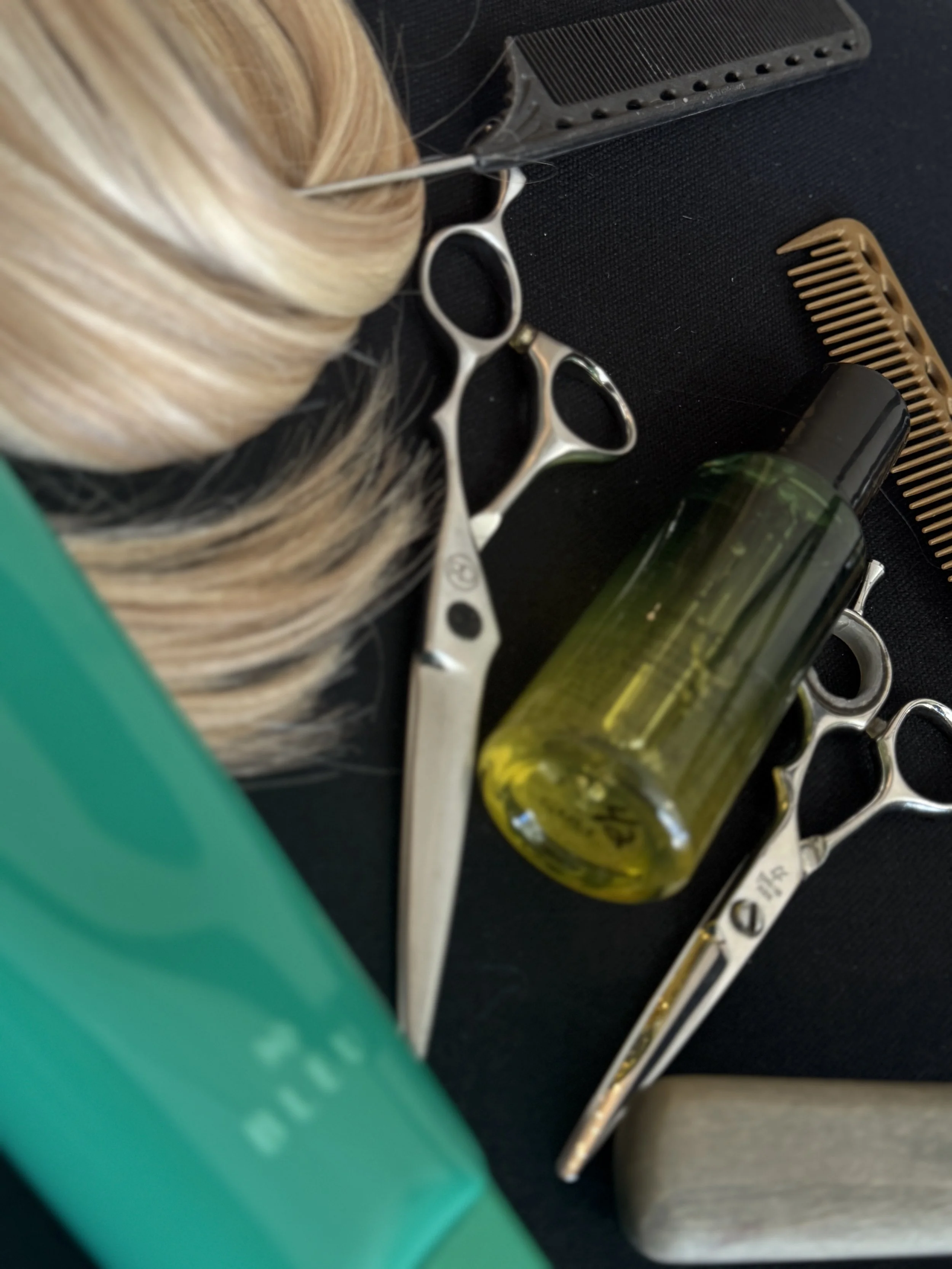 Tools on a black surface, including a pair of silver hairdressing shears, a green bottle with a black cap, a hairbrush, a brown hair clip, a black comb, and a gray rectangular container.