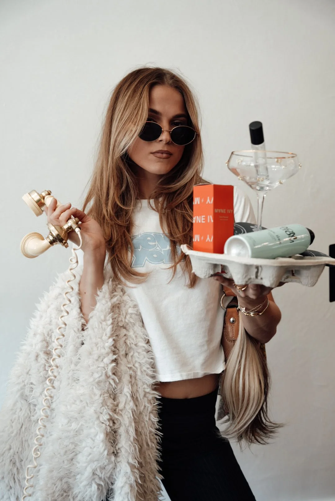 A young woman with long blonde hair wearing sunglasses, a white graphic t-shirt, and a fuzzy jacket, holding a vintage beige telephone in one hand and a tray with a cocktail glass, a red box, and a can in the other.