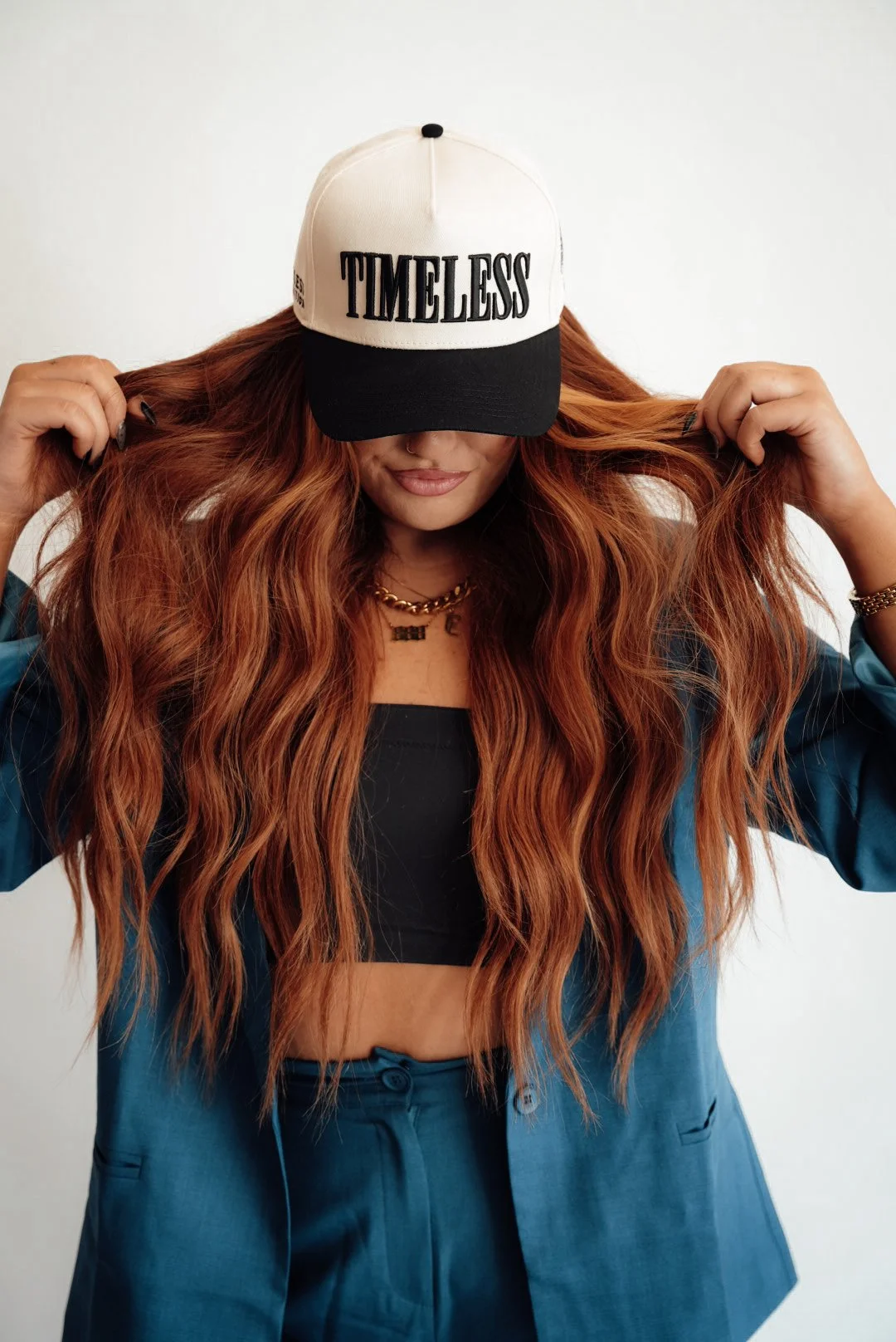 A woman with long, wavy red hair wearing a white and black baseball cap with the word "TIMELESS" on it, a black crop top, a blue blazer, and blue pants, holding her hair with both hands.