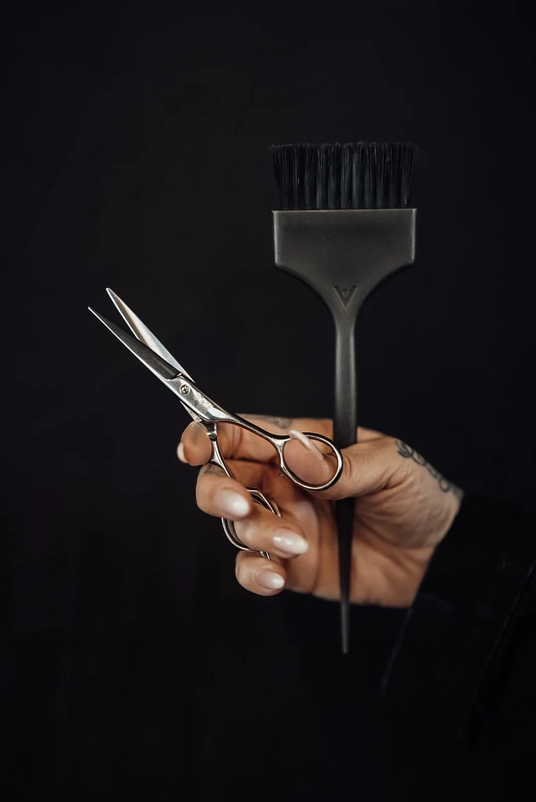 Hand holding hairdressing scissors and a hairbrush against a black background.