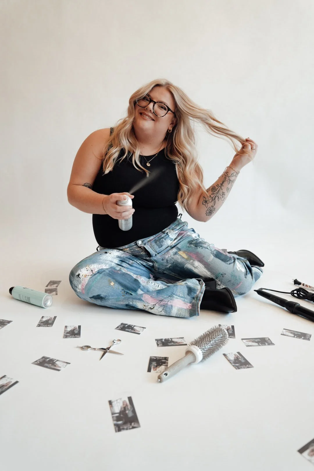 A woman sitting cross-legged on the floor, holding a spray bottle, with various hair and beauty tools around her, including a round brush, scissors, curling iron, and photos.