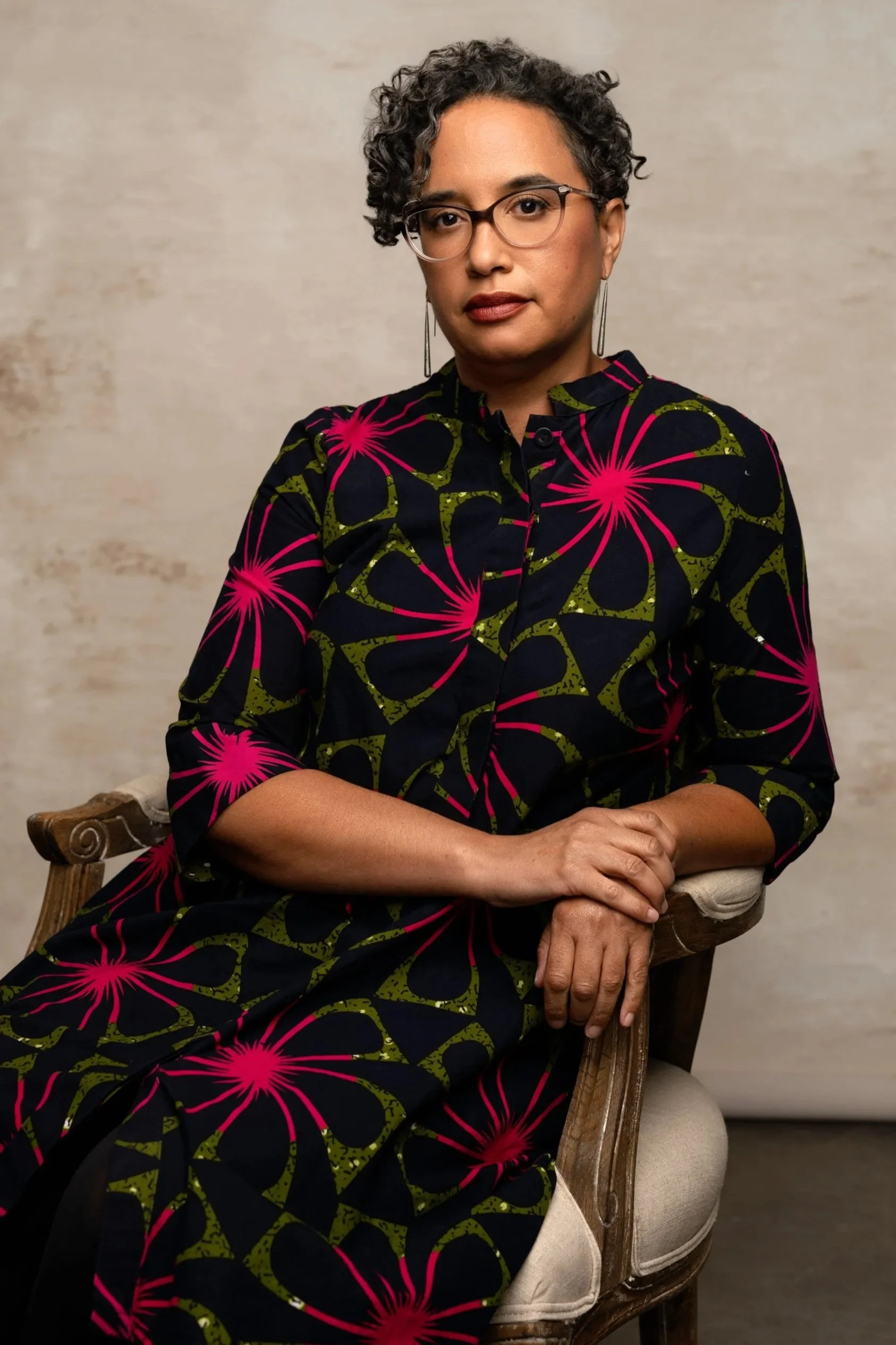 A woman with short curly hair, glasses, and earrings sitting on a wooden chair, wearing a black dress with pink and green floral patterns and looking at the camera.