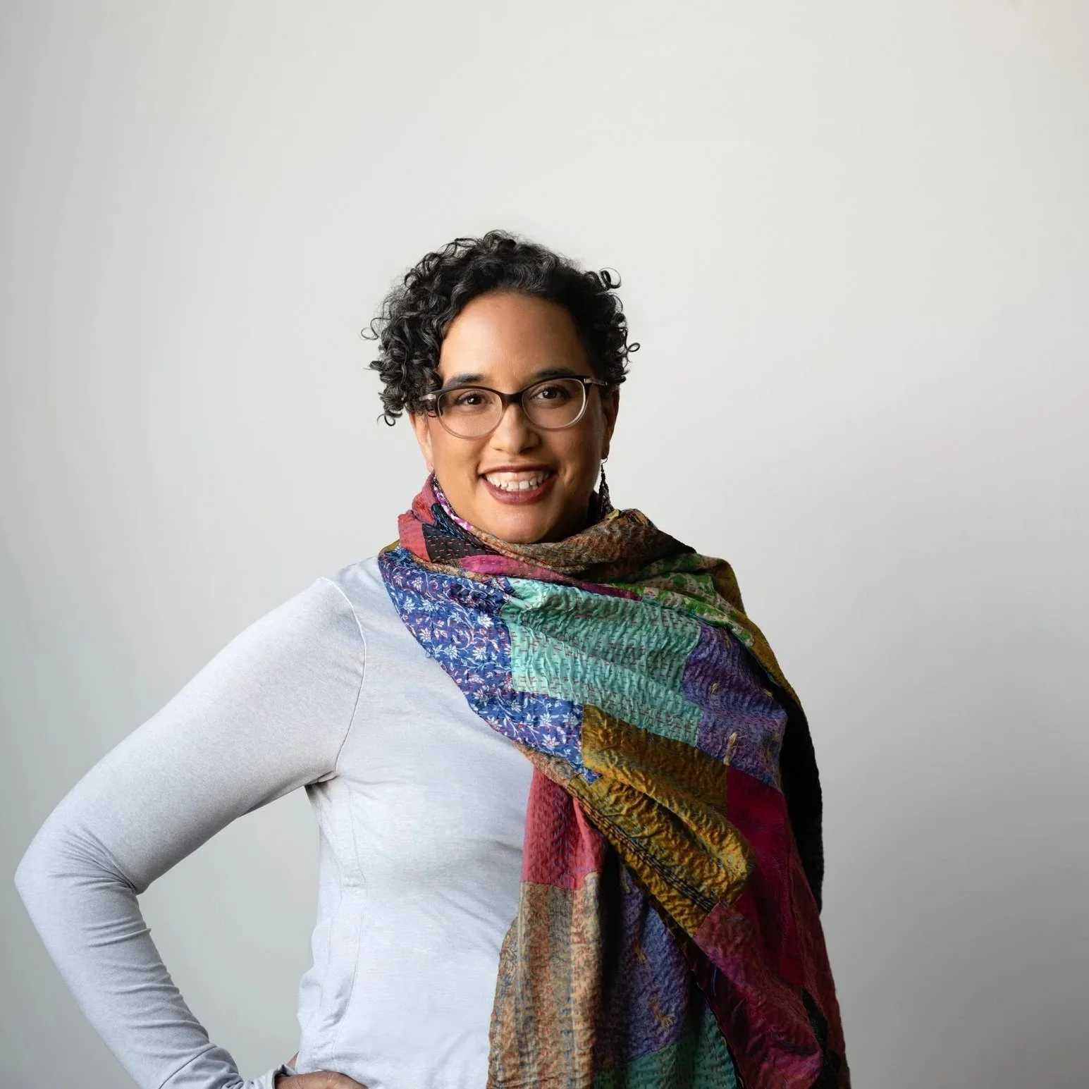 A woman with short curly dark hair and glasses, wearing a white long-sleeve shirt and a colorful patterned scarf, smiling and posing with her hand on her hip against a plain white background.