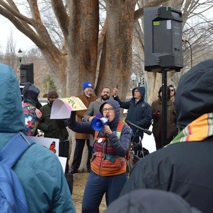 Shannon Gibney, a candidate for Minneapolis School Board (District 3) speaking into a megaphone at a gathering outdoors, surrounded by people in winter clothing. A speaker is mounted on a tall stand near her, and large trees are in the background.