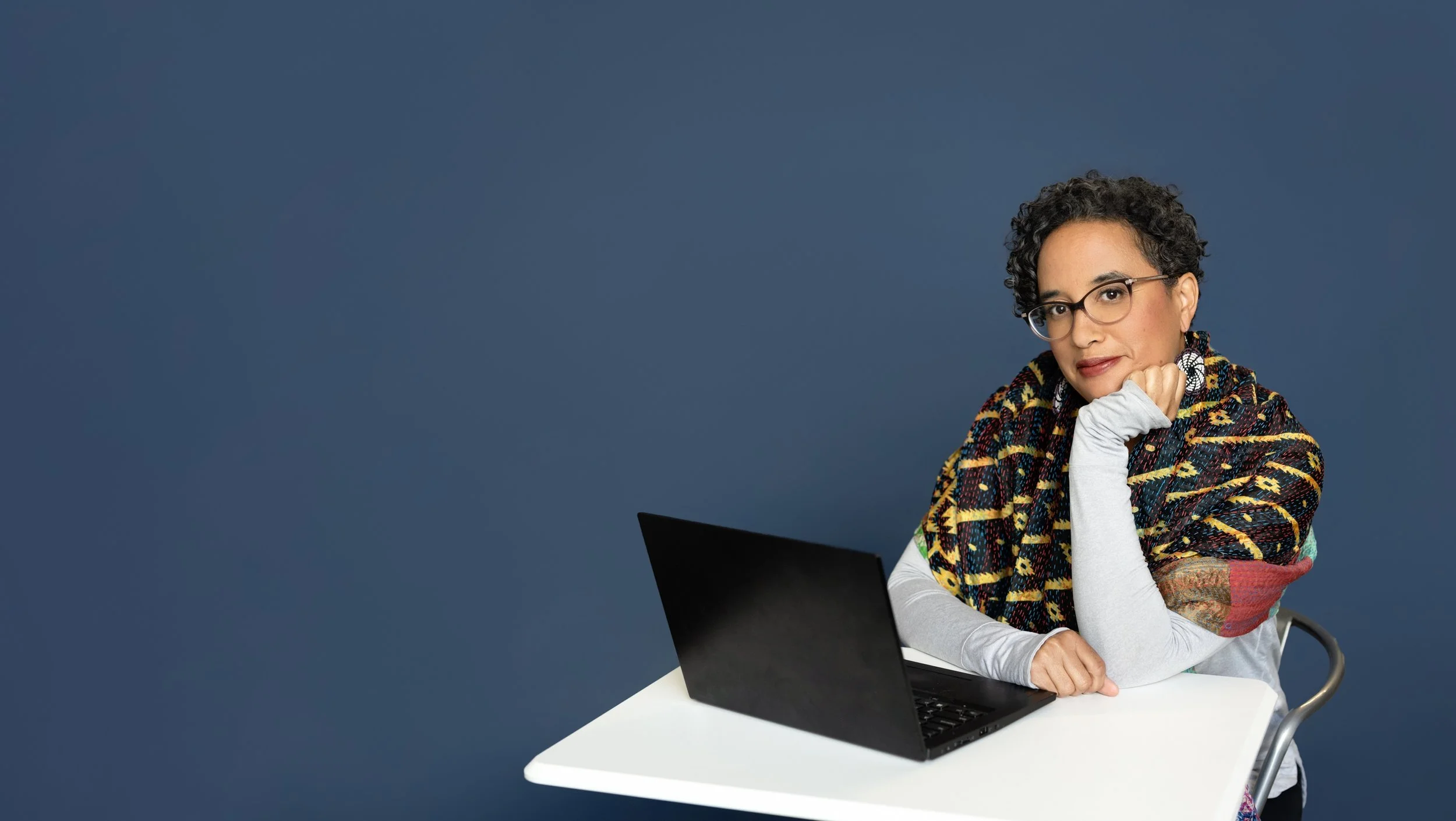A picture of Shannon Gibney, candidate for Minneapolis School Board, District 3. She has short curly hair, glasses, and colorful clothing sitting at a white table with a laptop, resting her chin on her hand, against a plain blue background.