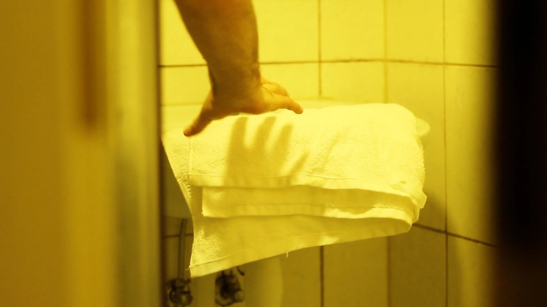 View of a person's arm and hand reaching out to touch a stack of white towels on a shelf, with tiled yellow walls surrounding the scene.