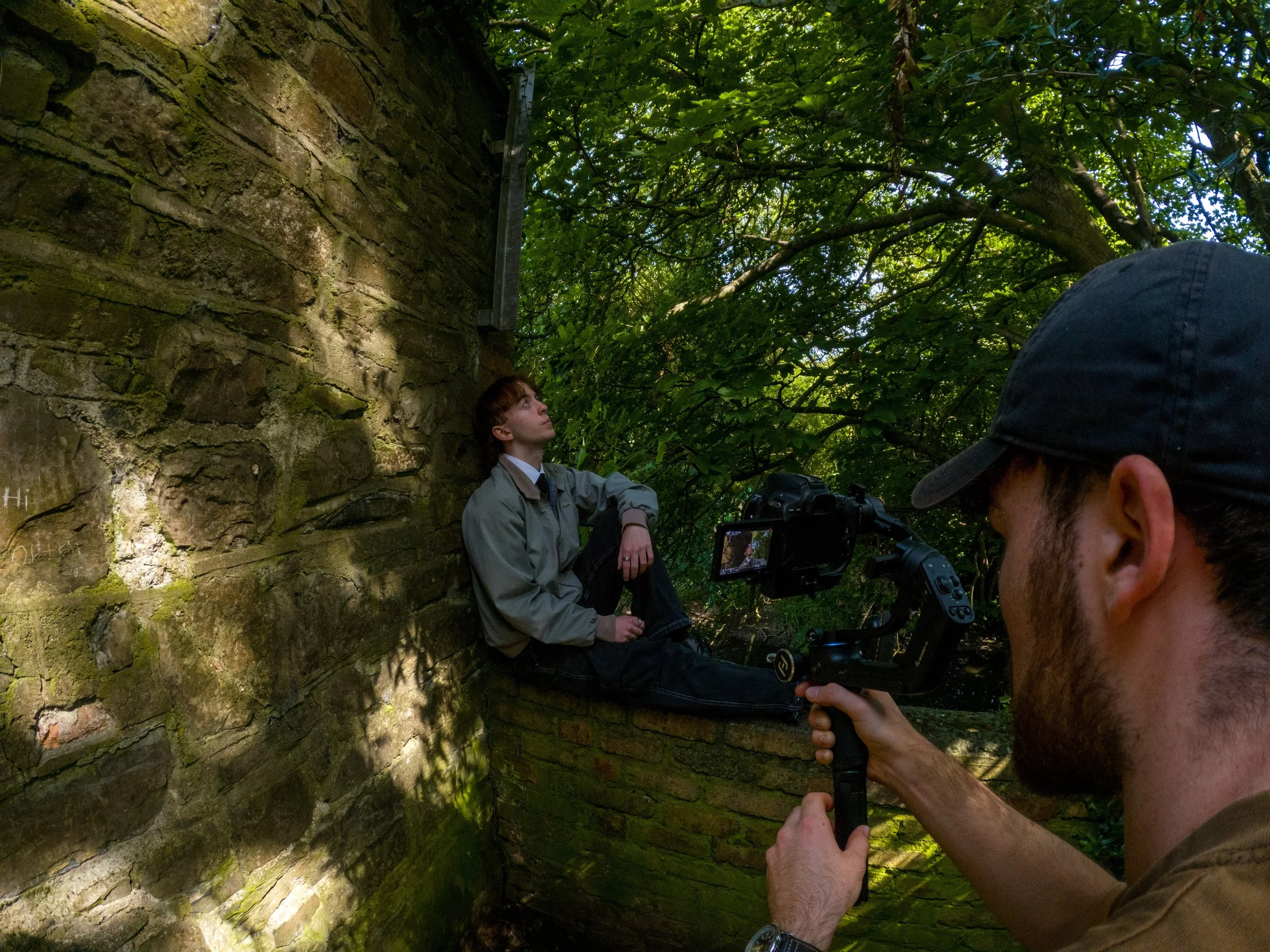 A woman is sitting next to a moss-covered brick wall amidst green trees, while a man in the foreground operates a camera that is focused on her.