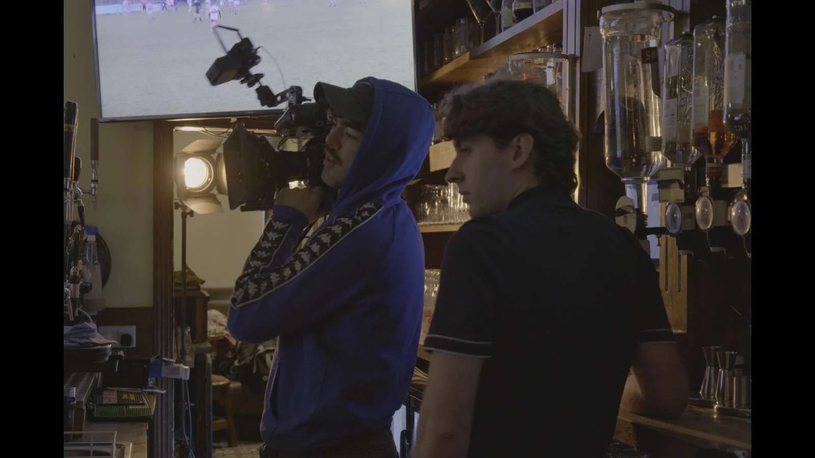 Behind the scenes of a video shoot showing a cameraman with a shoulder-mounted camera and a crew member at a bar, with filming equipment and glasses in the background.