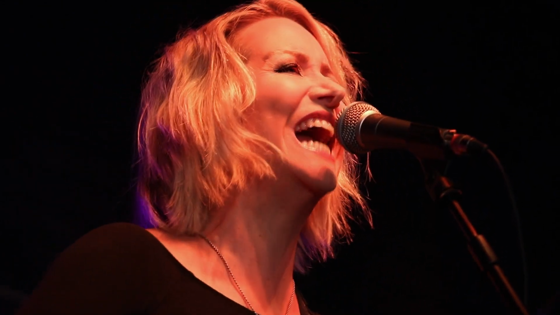 A woman with blonde, curly hair singing into a microphone on a dark stage.
