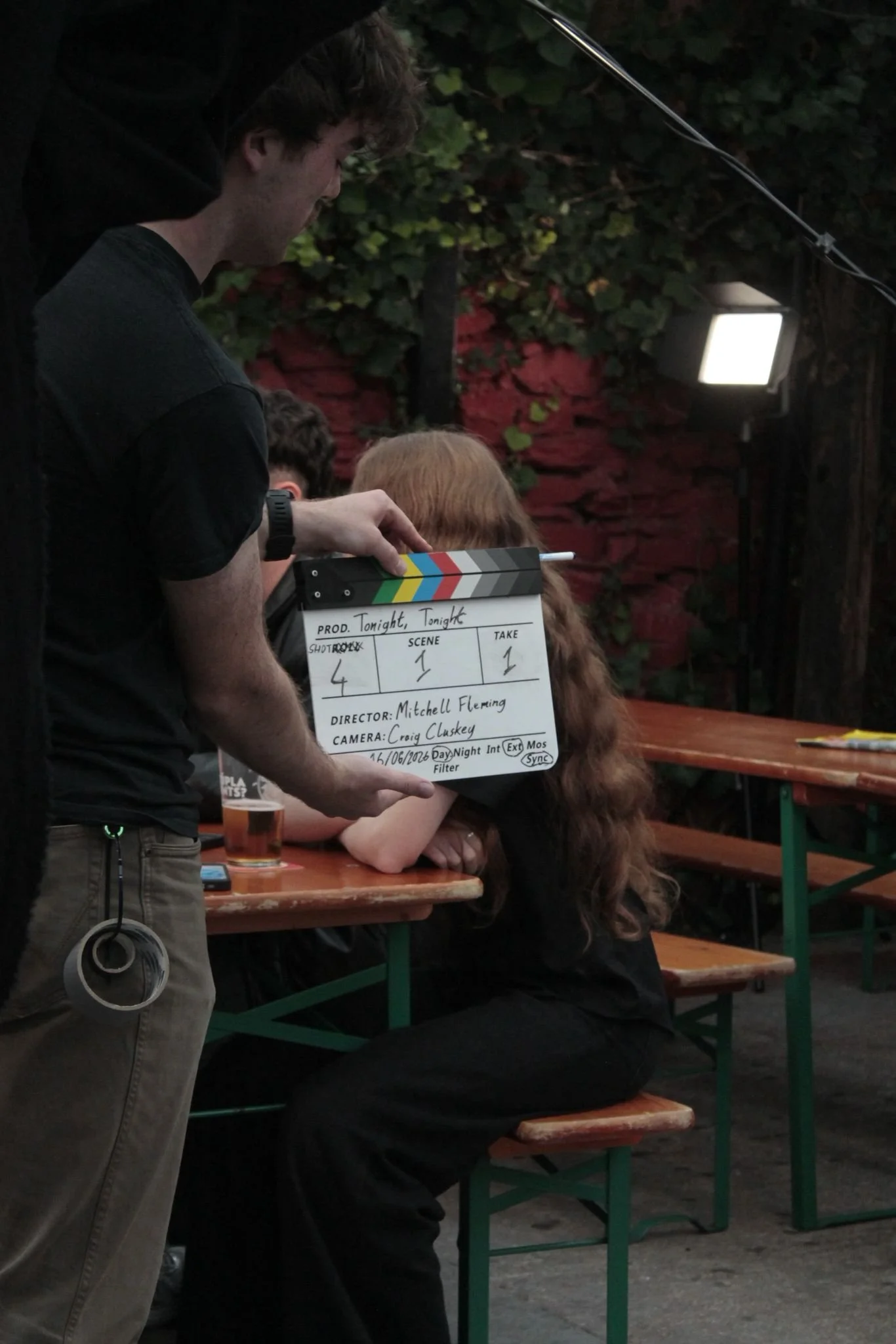 A person holding a film slate, standing next to a country-style wooden picnic table with people seated. The scene is outdoors under a canopy of green ivy. The slate has production details written on it, including the title 'Tonight, Tonight' and the director's name 'Mitchell Fleming'.