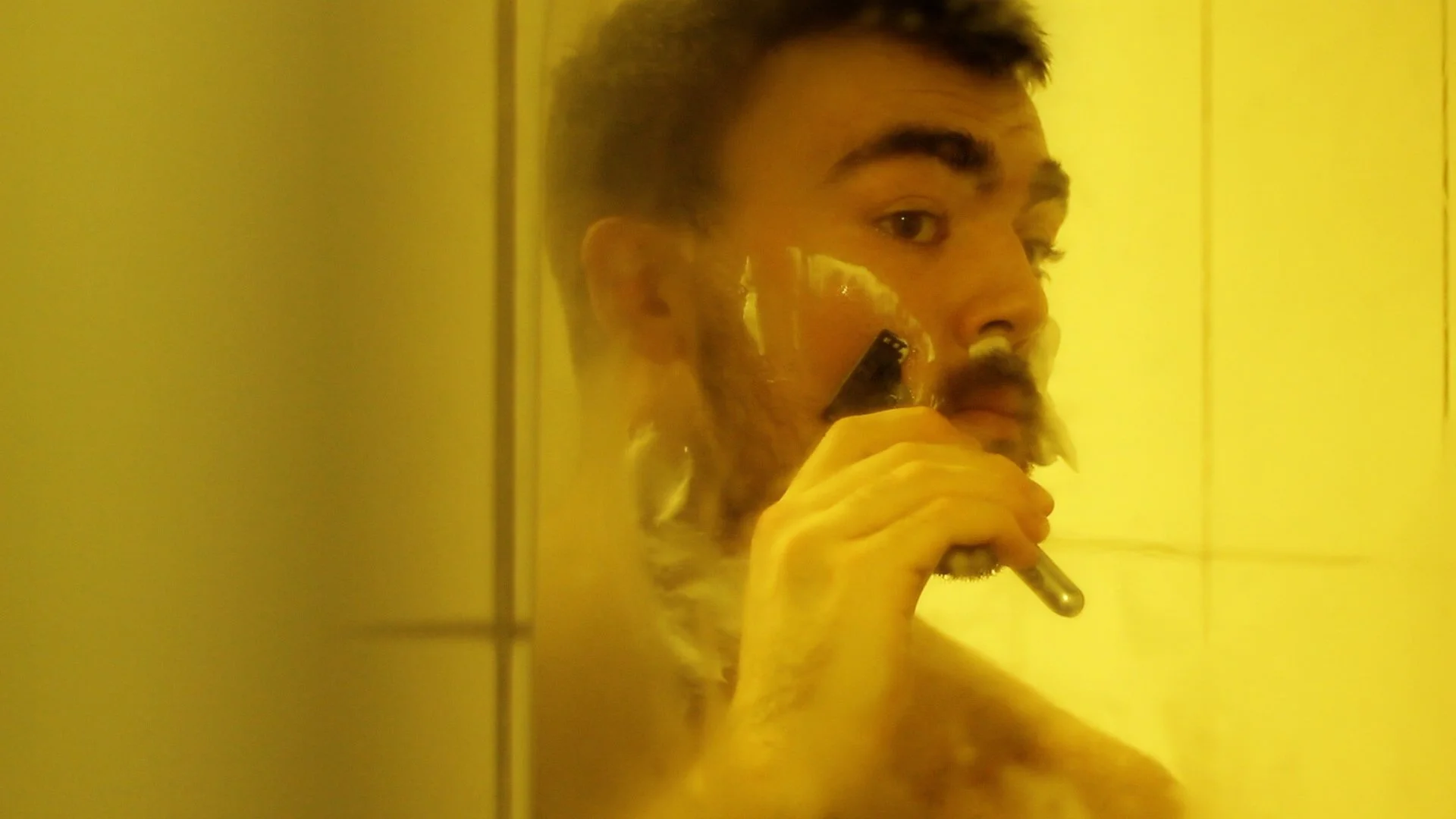 A person with short hair and a beard shaving their face in front of a mirror, holding a razor and using shaving cream.