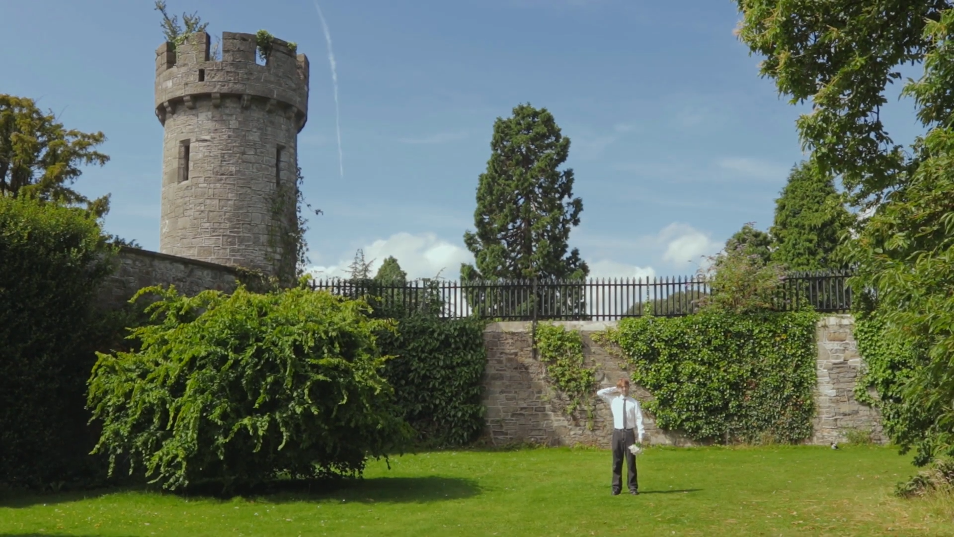 A man in a white shirt and dark pants stands on a grass lawn, talking on the phone, with a backdrop of lush greenery, stone wall, and a tall stone castle tower under a blue sky.
