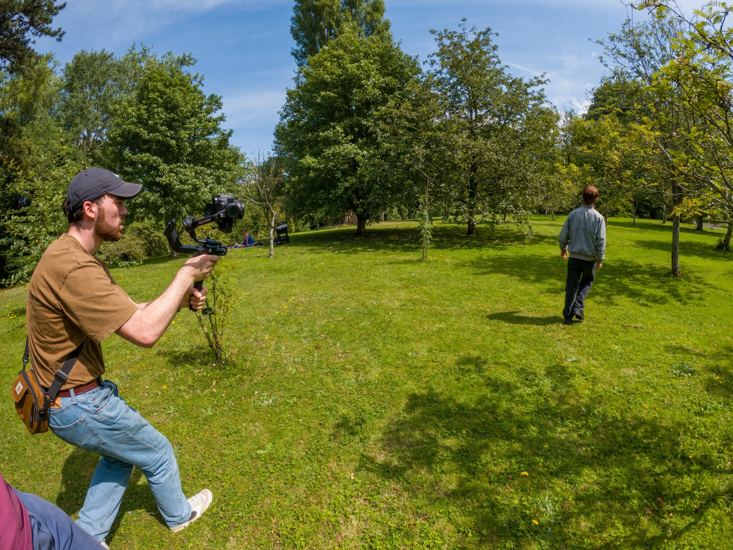 A person holding a camera stabilizer, another person walking away, and in the background they are sitting on the grass among trees on a sunny day.