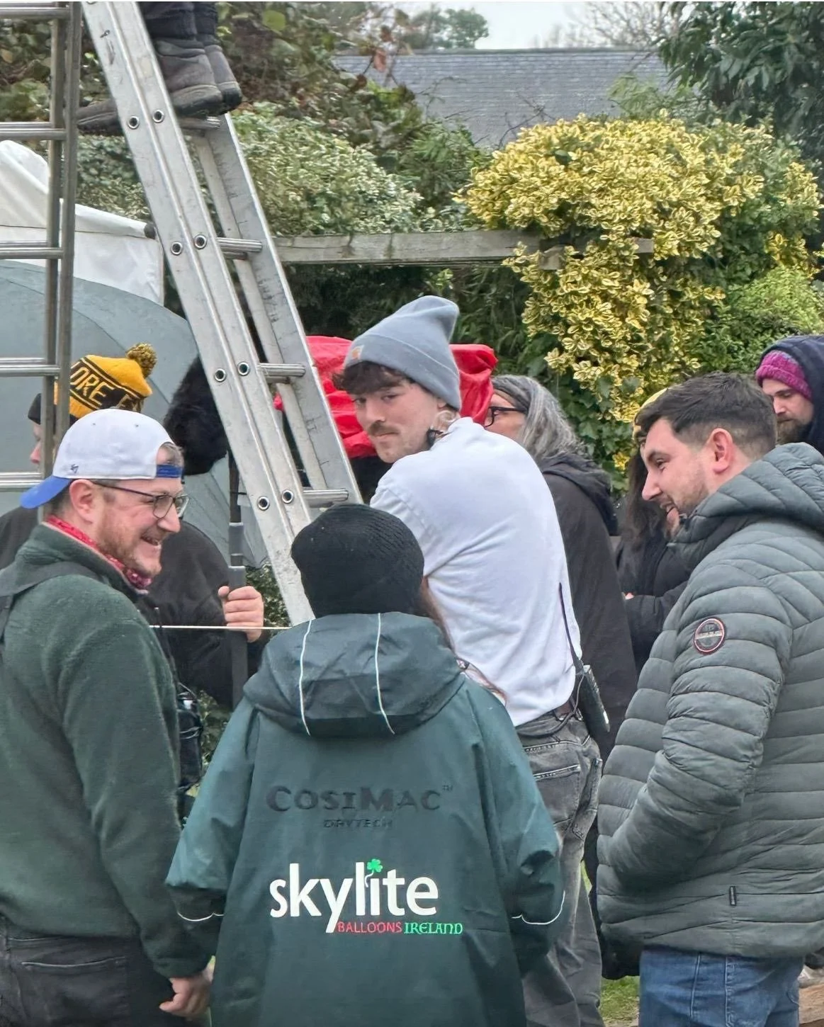 Group of people gathered outdoors around a ladder, with some smiling and wearing winter clothing, in front of trees and a house.