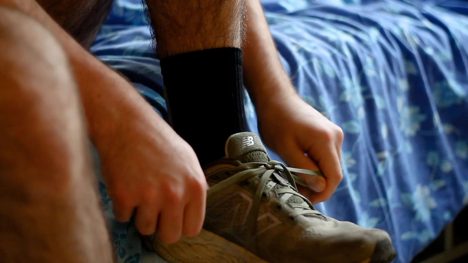 A person tying shoelaces on sneakers, wearing a black sock, with a blue floral-patterned bedspread in the background.