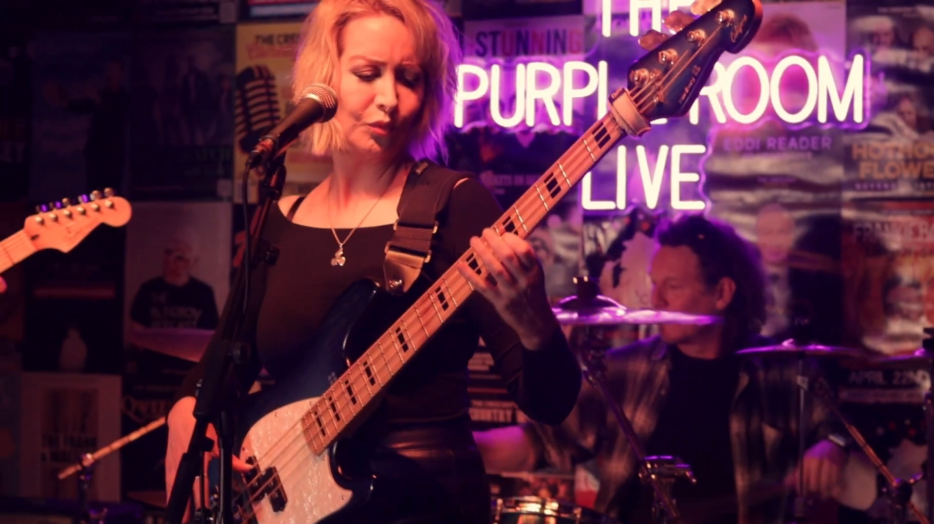 A woman playing bass guitar and singing into a microphone at a bar or music venue with neon signs, including a pink neon sign that reads 'Purple Room Live' and posters on the walls.