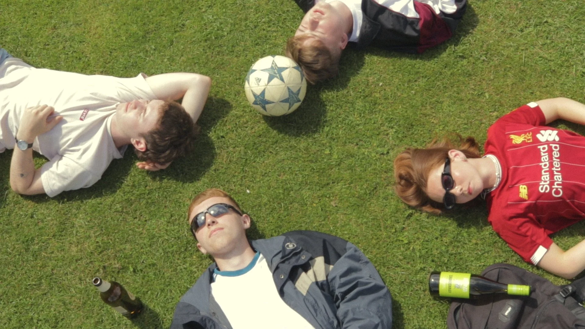Group of five young adults lying on grass in a circle with a soccer ball at the center.