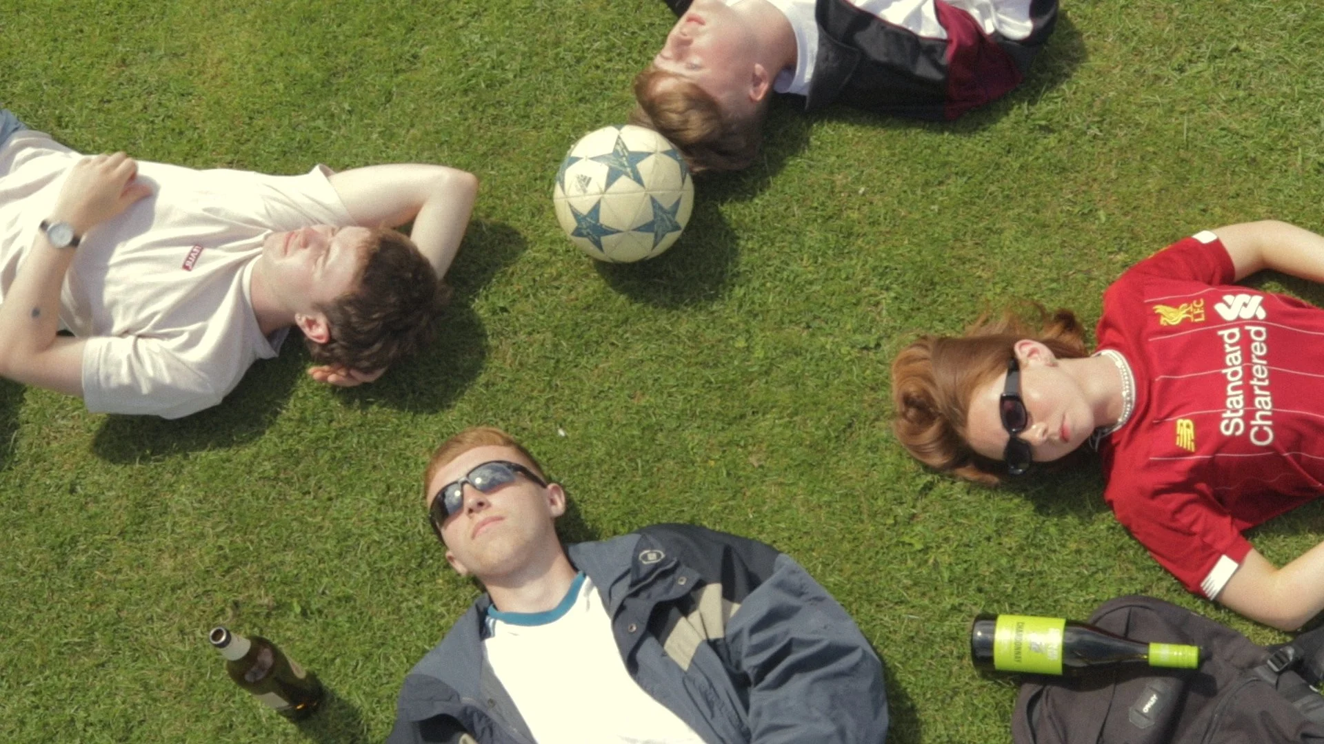 Group of friends lying on the grass in a circle with a soccer ball in the middle, some wearing sunglasses and casual clothing.