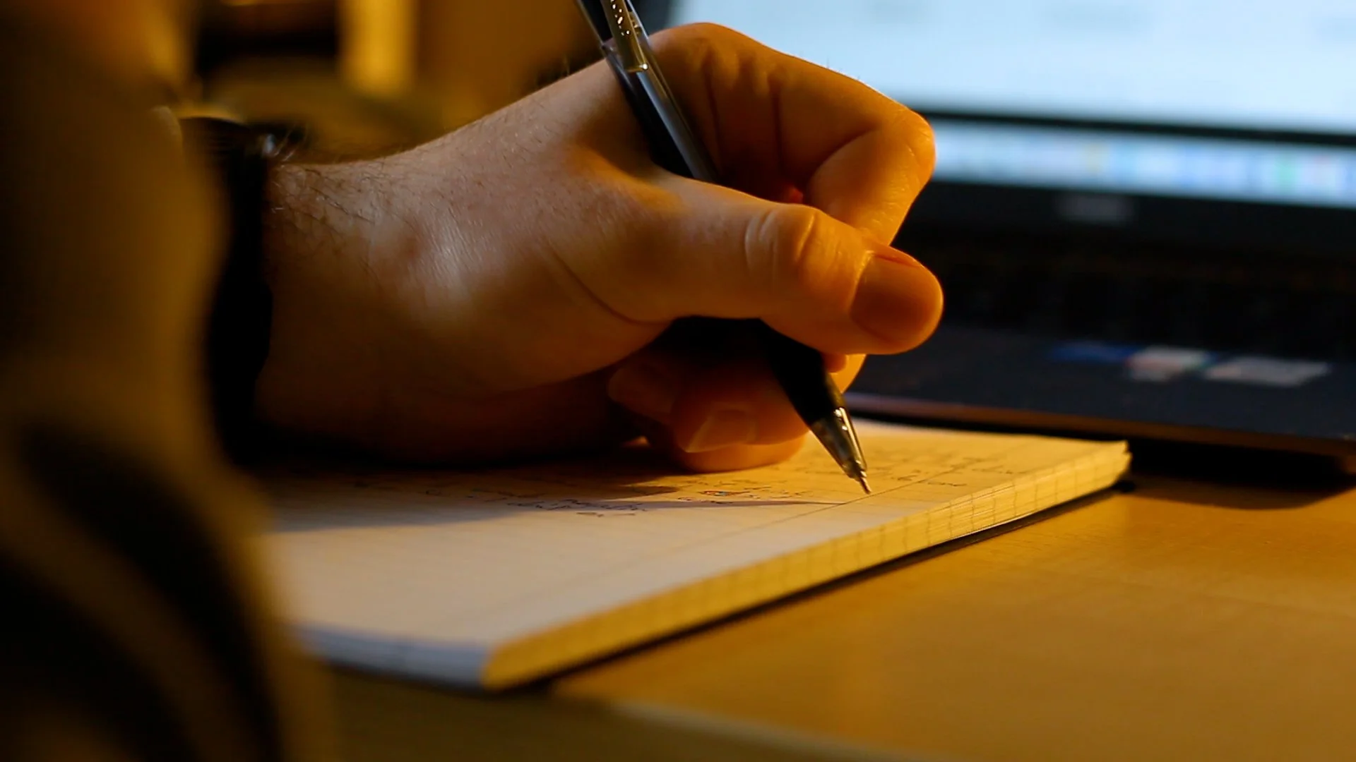 Close-up of a person's hand holding a pen and writing on a notepad with a computer screen in the background.