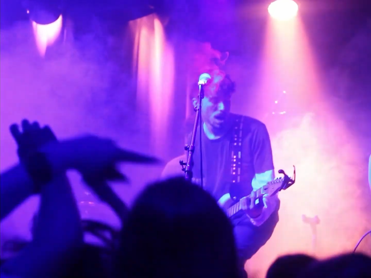 Musician playing guitar on stage under pink and purple lights, with a microphone stand in front and audience hands in the foreground.