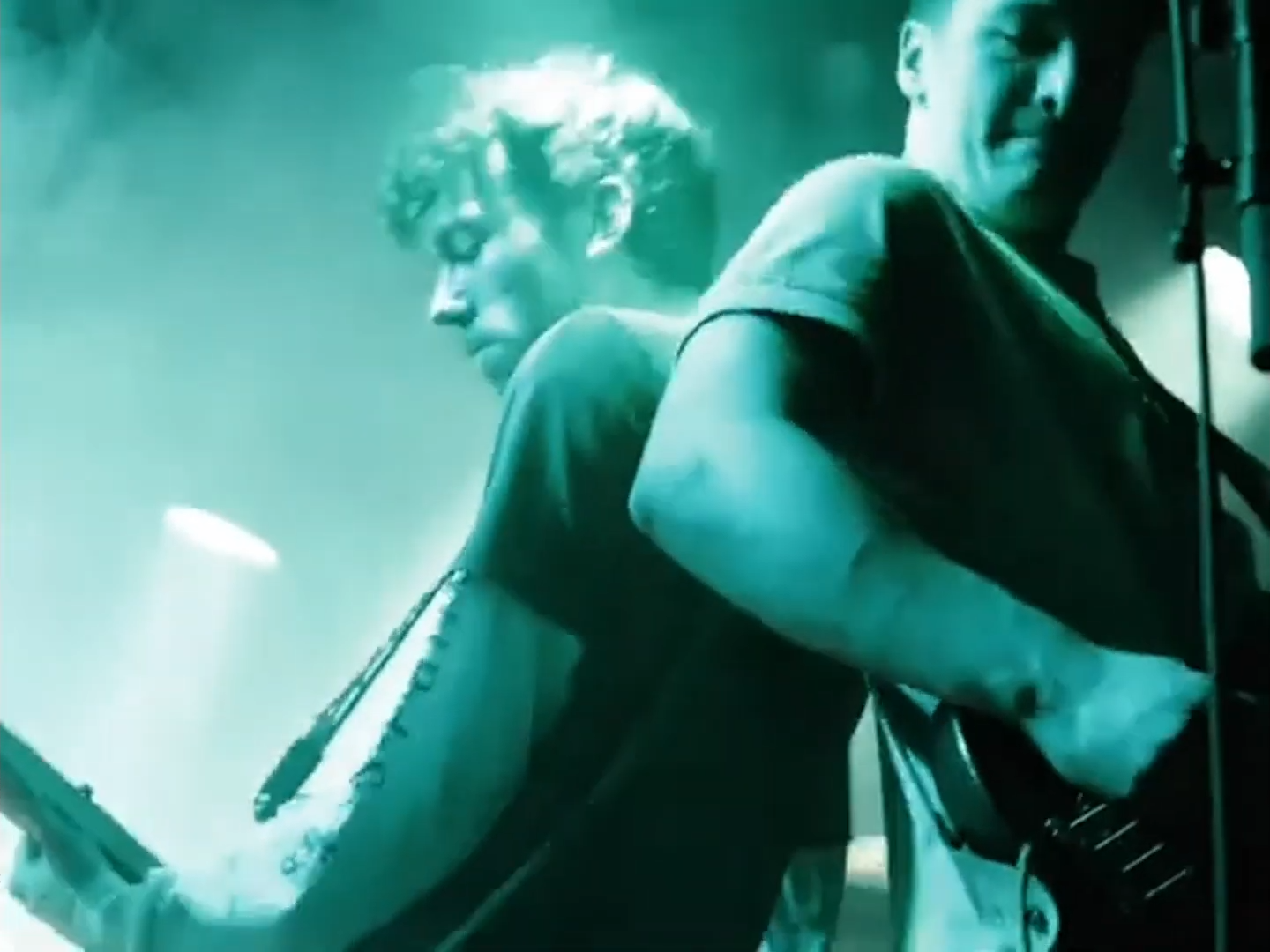 Two musicians perform on stage; one playing keyboard and the other singing or playing guitar, with a blue-green lighting ambiance.