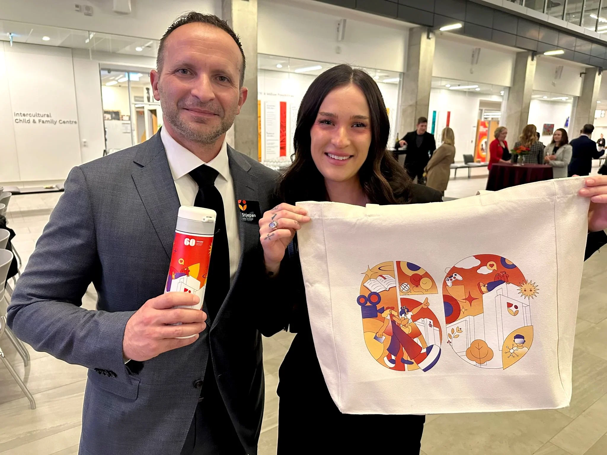 A man in a gray suit and a woman in a black dress holding a tote bag with a colorful design, standing inside a modern building with people in the background.