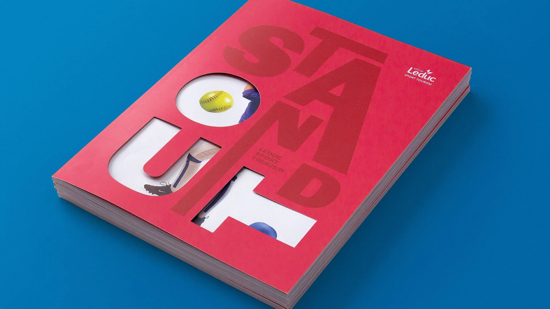 Red brochure with cutouts showing images related to sport and tourism, including a yellow softball, an athlete's legs, and a blue ball, placed on a blue surface.