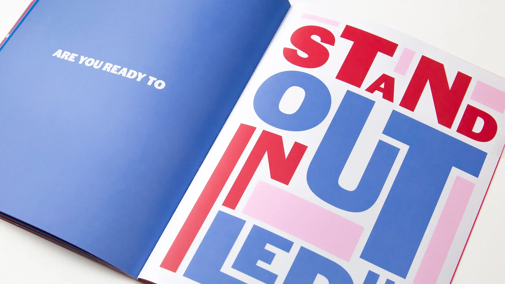 Open booklet with colorful text on the pages. The left page is blue with white text reading "ARE YOU READY TO". The right page has large, bold, red and blue text saying "STAND OUT IN THE CROWD" with some pink highlights.